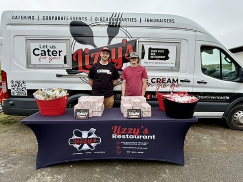 Boxed lunches and drinks on a Lizzy’s catering table in front of the branded van at a golf event